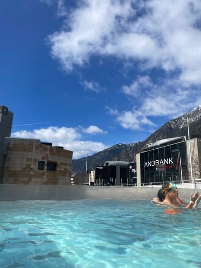 A swimmer wearing goggles and an orange top is in a pool of clear blue water. The location is likely the city of Les Escaldes, Andorra, as evidenced by the prominent "ANDBANK" building in the background. The building, with modern glass architecture, features the bank's logo. The foreground shows the pool's edge. In the distance is a partial view of mountains, with snow visible on the higher slopes, and a bright blue sky with scattered white clouds. Other buildings are also visible, including a beige-tiled structure.