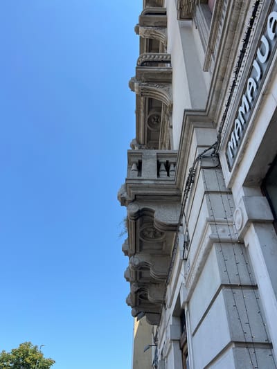 The image shows an architectural detail of a building in Bari, Italy, with a clear blue sky. The building features ornate stone detailing, including balconies with decorative moldings and classical architectural elements. White lettering, partially visible, spells out "MAMAJU..." and appears to be part of a business sign on the building's facade. String lights with small bulbs are draped along the architectural trim. A tree with green foliage is visible in the lower left corner of the frame, and a street lamp is also present. The perspective is looking up towards the building.