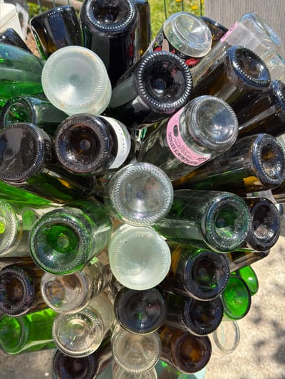 The image shows a cluster of empty wine bottles, predominantly green and brown, viewed from the top. The bottles are arranged tightly together, showcasing their bottoms and necks. Some bottles have labels still attached. The setting appears to be outdoors, with a glimpse of a rough surface, possibly concrete or stone, visible in the background. Felgueiras, Portugal, is not discernible from the visual information provided.
