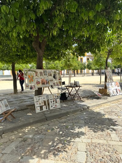 This image captures an outdoor art market scene in Sevilla, Spain. The setting is a sunny, cobblestone street or plaza, shaded by numerous lush green trees, identifiable as orange trees due to the foliage and depicted fruit in some artworks.
In the foreground and midground, several white display stands are set up, showcasing various framed artworks, likely prints or paintings. One prominent artwork on the larger stand depicts oranges on a branch. Other pieces feature diverse subjects, including botanical illustrations and stylized figures. A person, appearing to be a woman dressed in a red top, blue jeans, and white sneakers, stands near the largest display, looking down, possibly at her phone or at the artworks.
The ground is paved with irregular cobblestones and large square stone slabs, with strong, dappled shadows from the trees indicating bright daytime conditions. In the background, traditional white buildings with distinctive yellow trim and dark windows are visible, characteristic of Andalusian architecture. Folding chairs and additional display tables are also scattered around, suggesting a vendor's setup. A small black sign on one of the smaller stands clearly displays the number "15," likely indicating a price. The overall atmosphere suggests a relaxed, leisurely day in a historic European city.