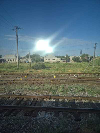 The image depicts a sunny day in Turkistan, Kazakhstan, likely from a moving train. In the foreground, multiple sets of train tracks run horizontally across the frame, covered with ballast and wooden sleepers. Beyond the tracks, a grassy field stretches towards a line of houses with tiled roofs. Power lines and utility poles are visible throughout the scene, supporting the electrical infrastructure. The sky is bright blue with a strong sunflare obscuring part of the upper right quadrant. A blue tractor is parked in front of one of the houses.
