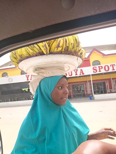 The image depicts a woman carrying a head-load of bananas in Hotoro, Nigeria, captured from inside a vehicle during daylight. The woman, shown from the chest up, is positioned outside the vehicle, with her face in profile looking towards the right side of the frame. A large, white, round basin filled with ripe, yellow bananas (some with brown spots) is balanced on her head, supported by a light-colored fabric pad. She wears a teal-colored hijab covering her head and shoulders. A small nose ring is visible in her right nostril, and her mouth is slightly open. Her right arm and hand are partially visible in the lower right foreground, appearing within the vehicle's interior frame. In the background, a commercial building with a yellow and red facade and white trim is prominent, featuring a sign with large red lettering that reads "SUYA SPOT". The immediate ground appears paved.