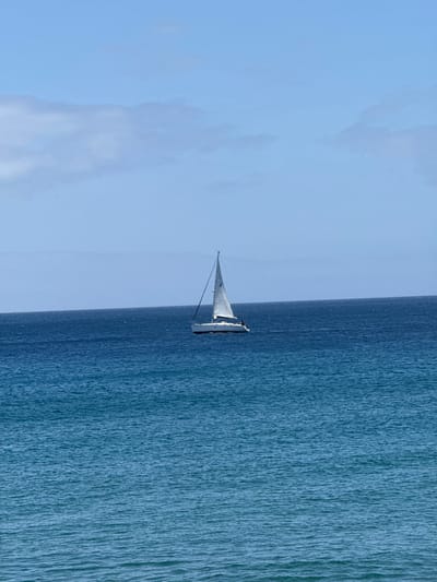 A serene maritime scene captures a lone sailboat gracefully traversing the expansive blue waters under a bright, clear sky. The image, when oriented correctly, shows a distinct horizon line separating the calm ocean from the vast expanse of the sky.
In the mid-ground, a white-hulled sailboat with a triangular white mainsail and a jib is the central subject. The sails are unfurled, indicating that the boat is actively sailing. Details of the mast, rigging, and parts of the deck are visible. The boat appears to be sailing across the frame, creating a sense of tranquil movement.
The ocean dominates the lower half of the scene, showcasing a deep, rich blue color. The water's surface is relatively calm, with small, textured ripples and subtle waves reflecting the daylight. The clarity of the water suggests a pristine marine environment. Above the horizon, the sky fills the upper half of the image with a lighter, ethereal blue. A few faint, wispy clouds are subtly present, adding depth without overshadowing the overall brightness of the day.
The lighting suggests it is daytime, likely midday or early afternoon, with abundant natural light illuminating the scene. The weather appears ideal for sailing, characterized by clear skies and calm seas. The setting evokes feelings of peace, freedom, and the quiet majesty of nature. This picturesque moment unfolds off the coast of Puerto Del Carmen, Spain, highlighting the beautiful coastal waters of the region. There is no visible text in the image.