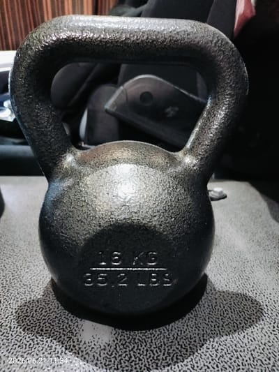 A kettlebell with "16 KG" and "35.2 LBS" embossed on its base sits on a textured grey surface. The kettlebell is dark, with a speckled finish, and has a rounded body and a thick, curved handle. The background is out of focus, showing dark shapes that appear to be bags or gym equipment, with a hint of a red and white object. The lighting is somewhat dim, casting a shadow from the kettlebell onto the surface. The timestamp "2026.06.21 11:34" is visible in the bottom left corner of the image. The location of Gorizia, Italy, is not discernible from the image content.