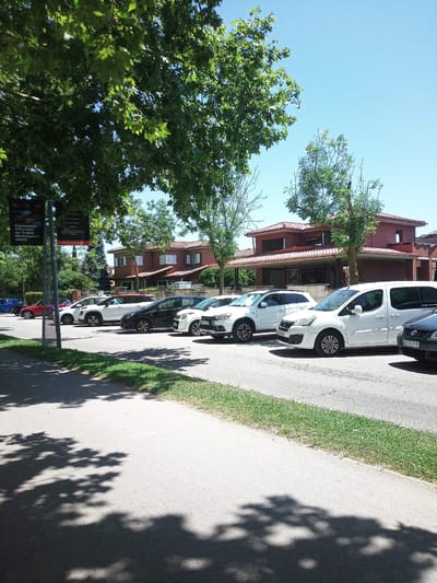 This media file depicts an outdoor scene on a sunny day, likely in the late morning or early afternoon, given the bright light and the sharp shadows cast by the trees. The setting appears to be a residential area or a public space with parking. A row of cars is parked along the side of a paved road. Among the vehicles are a white minivan, a white SUV, a dark colored sedan, a red hatchback, and a white compact car. In the background, a two-story building with a red tile roof and light-colored walls is visible, surrounded by trees and greenery. Some of the trees have lush green foliage, and the sky is a clear, bright blue. A signpost with informational banners is also present near the left side of the frame, partially obscured by tree branches. The banners are black with white text, and one of them reads "Campionat d'Europa de Triatló Multiesport 2020," suggesting the location is Banyoles, Spain, and the event referenced is the European Triathlon Multisport Championships in 2020. The scene is peaceful and evokes a sense of a typical day in a suburban or town environment during warm weather. There are no people visible in the frame.