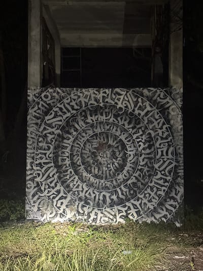 The image depicts a circular mural of calligraphy painted on a dark background, possibly a wall or panel. The calligraphy is intricate and abstract, rendered in shades of white and grey, creating a visually complex pattern that radiates outwards from the center. To the left of the mural, there is a dimly lit interior space with concrete structures, suggesting an abandoned or unfinished building. A doorway or opening leads into the dark interior, and some debris is visible on the floor. To the right of the mural, a patch of dry, green grass is illuminated by a light source, possibly a flashlight or street lamp, casting a harsh glow. The overall lighting suggests it is nighttime or a very dark environment. The presence of grass and the hint of a tropical environment (given the location context) are the only clues to the outdoor setting. There is no clear activity or event taking place; the focus is on the static artwork and its surroundings. No people are visible. The calligraphy itself is abstract and does not appear to form discernible words in a common language. The location context indicates Ko Pha-ngan District, Thailand, though this is not visually evident within the image itself beyond the potential for lush vegetation.
