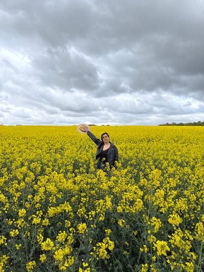 This image captures a vibrant yellow rapeseed field under an expansive, cloudy sky. The field, densely packed with bright yellow flowers, stretches from the foreground towards a distant line of dark green trees that mark the horizon.
A young woman is positioned in the lower right section of the frame, kneeling or crouching amidst the yellow blossoms. She is dressed in a dark jacket, possibly leather, over a dark top, and holds a light-colored, wide-brimmed hat in her outstretched right hand. Her dark hair is visible, and she looks towards the camera with a subtle smile, conveying a sense of calm and enjoyment.
The sky is dominated by heavy, grey, and white clouds, suggesting an overcast day. This weather creates a diffused light across the scene, highlighting the brilliant yellow of the flowers against the more muted tones of the sky. The scene depicts a peaceful moment within a rural, agricultural landscape, consistent with locations like the Breckland District, United Kingdom, known for its extensive fields of crops. No visible text is present in the image. The overall impression is one of a person posing and appreciating the beauty of nature.