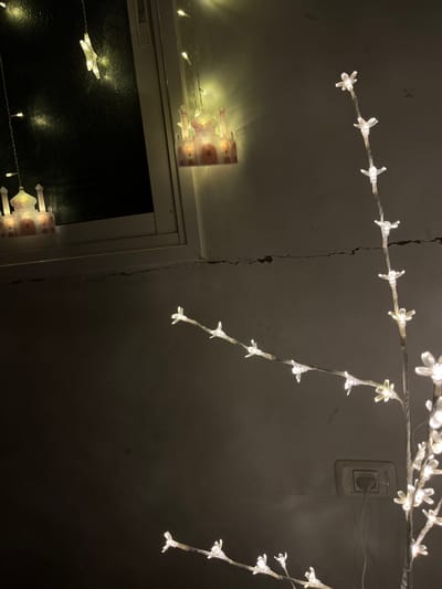 The image displays an indoor scene at night, likely within a residence in Nuseirat, Palestinian Territory, illuminated by decorative lights. In the upper left quadrant, a white-framed window with dark glass is visible, reflecting internal lights. String lights with warm yellow illumination are present; one mosque-shaped light is affixed to the wall beside the window frame, and another identical light is visible through the window. A star-shaped light is also seen near the top left of the window. The wall beneath the window exhibits prominent horizontal cracking. On the right side of the frame, a decorative light tree or branch, adorned with numerous small, translucent, white, flower-shaped LED lights emitting a bright white glow, extends vertically and horizontally. This light tree is plugged into a light-colored two-prong electrical outlet located on the wall in the lower right. The walls visible in the scene are a light, possibly off-white or beige color, and show additional horizontal cracking. No individuals or specific activities are depicted.