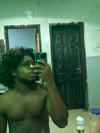 An interior scene captured in Gbongan, Nigeria, depicts a dark-skinned male taking a mirror selfie. The subject, shirtless and wearing a black headband, has dark, curly hair and a beard. A single gold stud earring is visible in his left ear. He holds a dark-colored smartphone in his right hand, positioned to capture his reflection.
The background, reflected in the mirror, shows walls with a light, possibly off-white, uneven texture. Numerous white flakes and dark streaks are present on the upper walls and ceiling line, indicating surface degradation or moisture damage. Two doorways are visible: one partially obscured on the left, showing an interior space with a faint reflection, and a prominent dark brown, paneled double door with a silver-colored handle on the right. In the lower right foreground of the reflection, the tops of two bottles are visible on a surface: a blue-capped white bottle and a tan bottle with a white pump dispenser. The entire image is dominated by a green color cast.