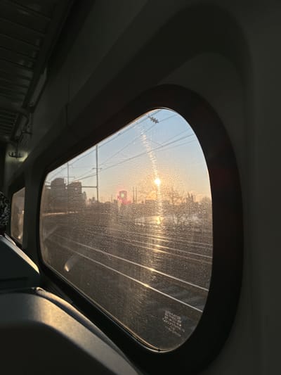 The image provides a forensic view from inside a passenger train, looking through a rounded rectangular window that is visibly coated with numerous small water droplets or particulate matter. The exterior scene is illuminated by a low sun, positioned in the upper-right, casting a bright glare and a shimmering golden reflection across a wide body of water, likely the Schuylkill River. Multiple parallel railway tracks, crossed by a network of overhead catenary wires, are visible in the mid-ground. A distant urban or industrial skyline, composed of dark building silhouettes, is present beyond the river, featuring a prominent red structure and a green railway signal. The interior of the train, largely in shadow, reveals a dark-colored seat partially illuminated by sunlight in the lower-left. The location is along the Schuylkill River in Philadelphia, United States of America. No individuals or specific actions are discernible within the frame.