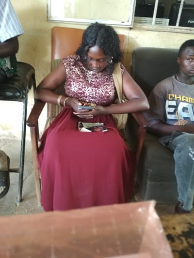 The image depicts an indoor scene, possibly a waiting area or a common room, in Bisichi, Nigeria. In the foreground, a woman is seated in a wooden chair, wearing a red dress with a patterned top. She is holding a mobile phone and appears to be engrossed in it. To her right, a man is seated on a dark sofa, wearing a grey t-shirt with text that appears to be "CHAMPIONS". He is looking towards the camera. Behind the woman, part of another person is visible, wearing a striped shirt. The environment appears somewhat sparse, with a wooden chair and a dark sofa as the main furniture. The background shows a wall and what might be a window or a framed picture. The overall impression is of a casual gathering or waiting period in a functional setting.