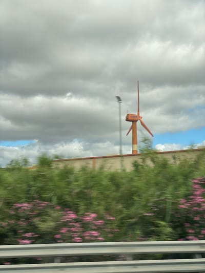 The image captures a close-up of a small orange wind turbine with two blades, positioned against a cloudy sky. To the right of the turbine, a building with a tan facade is visible, partially obscured by dense green foliage with scattered pink flowers. The scene is viewed from what appears to be a moving vehicle, suggested by the motion blur throughout the image, especially in the foliage and the sky. The weather appears overcast, with large, grey clouds dominating the sky, though a small patch of blue sky is visible between them. The turbine itself has some text visible on its housing, but it is illegible due to the image quality. The overall impression is one of a bright but cloudy day, with a sense of motion and a focus on renewable energy infrastructure within a natural and built environment.