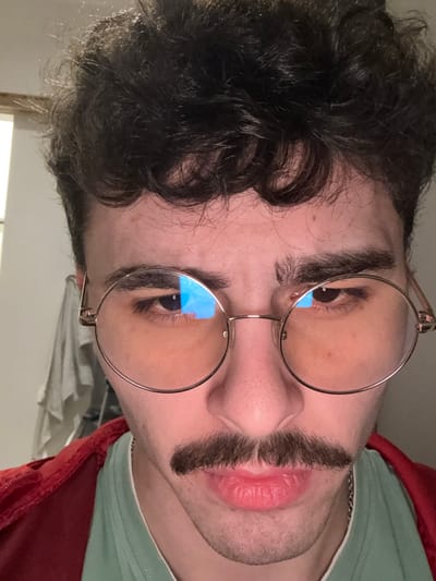 A person with dark, curly hair and round glasses poses for a photo. The image is shot from below, angling upward. The person's eyes are visible through the glasses, reflecting a bright blue light. A fake mustache is positioned between the nose and upper lip. The person wears a light green shirt with a red garment visible at the collar. A silver chain rests around the neck.