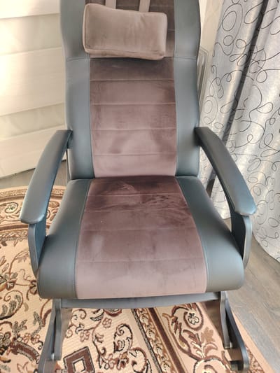 The image shows a close-up, eye-level view of a comfortable-looking, dark gray and brown upholstered chair, likely a gaming or executive chair. The chair has a high back with a separate headrest pillow, and wide armrests. The upholstery is a combination of smooth faux leather on the sides and outer frame, and a softer, velour-like fabric in horizontal bands on the back and seat. The chair is positioned on a patterned rug with ornate beige, brown, and black designs. Behind the chair, to the right, a portion of a gray curtain with a swirling black pattern is visible. To the left of the chair, a white door or wall with horizontal paneling is slightly illuminated. The lighting suggests an indoor setting, possibly daytime, with natural light contributing to the scene. The overall impression is one of comfort and relaxation, with no specific activity being depicted, just the chair as an object within a domestic or office setting. There is no visible text in the image. The location context is identified as Gorodets, Russia.