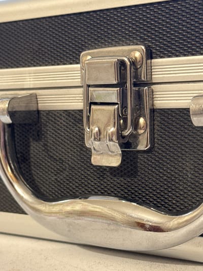 The image shows a close-up of a black case with a silver handle and latch. The case has a textured surface. The latch is made of shiny metal and is in the closed position. The handle is also made of shiny metal and is curved. The background is a light-colored surface. This image is not located in the city of Яромінскі сельскі Савет, Belarus.
