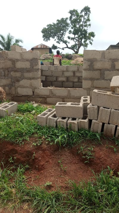 The image depicts an outdoor scene of a construction site in Kagarko South, Nigeria. A partially built structure, constructed from concrete blocks, dominates the foreground and midground. A man wearing a red and beige shirt and a black baseball cap is seated on the wall of the structure, facing away from the camera, looking towards a large tree in the background. Scattered concrete blocks are visible on the ground around the base of the partially constructed building, interspersed with green grass and red soil. In the background, a corrugated iron roofed building is visible, along with rocks and lush vegetation, including a prominent tree with spreading branches. The sky is overcast.