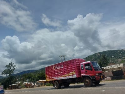 This image displays a red box truck, prominently branded with "e-posta" in white lettering on its cargo container, positioned on a paved road. The truck features a red cab and a large, red, horizontally ribbed cargo box with yellow/gold stripe accents, appearing to travel from left to right across the midground. The sky above is partially blue with numerous large, white and grey cumulus clouds. In the background, green, tree-covered hills with exposed rocky sections rise under the cloud line. To the left of the road, a series of small, makeshift structures or stalls with light-colored roofs are visible, interspersed with trees and green foliage. A large utility pole and overhead power lines traverse the upper portion of the frame. A small blue vehicle is partially visible at the bottom-left corner. No specific individuals or interactions are discernible. The scene is located in Kimamba, Tanzania.