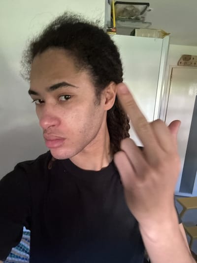 A man with dark curly hair and light skin is looking down at the camera, his right hand is raised towards his head with his middle finger extended. He is wearing a black t-shirt. The background is a white wall and a partial view of a window with a tan chair visible in the reflection. The location is unclear but presumed to be indoors, possibly a residence. No information about Les Ulis, France is discernible from the image.