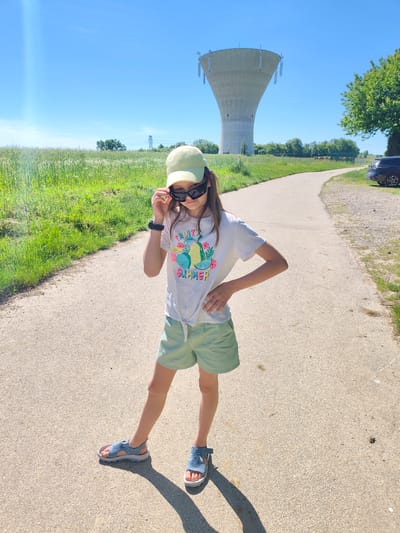 A young girl stands on a paved path, wearing a light green cap, sunglasses, a white T-shirt with the words "FRUIT SUMMER," light green shorts, and blue sandals. She holds the sunglasses on her face with her left hand and places her right hand on her hip. In the background, a water tower is visible, marking the location as Chaponnay, France. A grassy field is to the left of the path, and a road with a dark vehicle parked on the side is on the right. The sky is clear and blue.