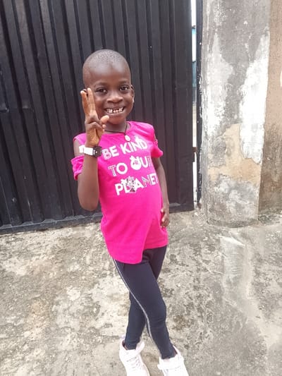 The image shows a young child standing outdoors in Igbede, Nigeria. The child has dark skin and a short buzz-cut hairstyle, smiling broadly with several gaps visible in the upper teeth. They are wearing a bright pink t-shirt with cut-out shoulders, which displays the white text "BE KIND TO OUR PLANET" and a graphic resembling Minnie Mouse. A dark necklace with a light-colored pendant is worn around the neck, and a white digital watch is present on the left wrist. The child is also wearing dark leggings with a thin light stripe down the side, and white athletic shoes secured with hook-and-loop fasteners. The child's left arm is raised, with the hand performing a two-finger peace gesture, while the right hand rests on the hip. The child's legs are crossed at the ankles. The ground beneath is grey, worn concrete. In the background to the left is a dark, vertically corrugated metal gate. To the right, a light-colored concrete pillar shows signs of wear and discoloration. A narrow opening between the gate and the pillar reveals a distant, indistinct structure.