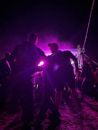 An outdoor nighttime scene is depicted at an event in Большая Кивара, Russia, characterized by strong purple artificial illumination. In the foreground, two individuals are largely silhouetted and appear to be in dynamic motion or dancing, facing each other. The person on the right is visible wearing a dark top and shorts, with their hair styled upwards, and arms in motion. Other silhouetted figures form a crowd in the mid-ground and background, indicating a public gathering. To the right, a metal truss structure, consistent with stage support or a sound system, is present. A prominent, thin pink line or cable extends diagonally from the upper right of the frame. The ground surface is unpaved, possibly dirt or sparse grass, reflecting the purple light. A atmospheric haze or smoke is visible throughout the illuminated areas.