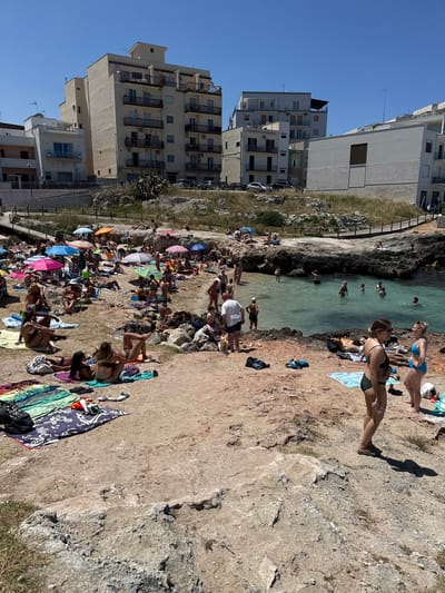 This image depicts a crowded beach cove in Monopoli, Italy, under a clear, bright blue sky. The scene is dominated by numerous individuals engaged in leisure activities on a sunny day.

In the foreground and mid-ground, a U-shaped cove with light turquoise-green water is visible. The shoreline consists of a mix of reddish-brown sand and grey, weathered rock formations. A significant portion of the sandy beach area on the left is densely populated with people. Many individuals are sitting or lying on colorful beach towels directly on the sand or rocks, some are under an array of vibrant beach umbrellas in shades of pink, blue, orange, red, green, and striped patterns. Activities on the beach include sunbathing, conversing, and walking. Notable objects include beach bags, sandals, and personal belongings scattered on towels.

Within the water of the cove, approximately a dozen individuals are visible. They are swimming, wading, or standing in the shallow water near the rocky edges. On the far right foreground, two women in bikinis are standing and conversing on a flat rocky outcrop. One woman wears a dark green bikini, and the other wears a light blue bikini. In the mid-ground, a man in a white t-shirt and dark shorts stands at the water's edge, facing away from the camera.

The background features an urban landscape. Directly behind the beach and rising uphill, there is an area of dry, sparse vegetation and light-colored rocky terrain. A wooden fence runs along a path or elevated walkway above the beach. Beyond this, several multi-story buildings of varying architectural styles and colors are visible. These appear to be residential or apartment buildings, featuring numerous balconies and windows. Buildings range from beige to white and light grey. Cars are parked along a road visible in the upper background, indicating a street level above the beach area. The overall impression is a bustling coastal urban environment.