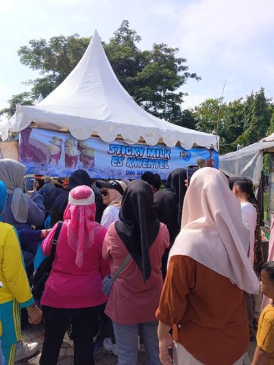 This outdoor image, taken in Ciamis, Indonesia, depicts a bustling crowd of people, primarily women, gathered around a food stall under a white, pointed tent on a sunny day. Many individuals in the foreground and midground are seen from behind, wearing various colored headscarves (hijabs) and casual attire, including pink, black, brown, and light beige.

A large banner stretched across the front of the tent advertises the stall's offerings. The banner features an image of several drinks in clear cups, topped with a white, frothy ingredient. Prominently displayed text in blue, bubbly letters reads "STICKY MILK ES KWENTEL" followed by "1DK." Below this, partially obscured by the crowd, the text "OM BE..." is visible.

The people appear to be queuing or congregating, with several looking towards the stall. One person in a light beige hijab and an orange-brown top is holding up a smartphone, possibly taking a picture or video of the scene. Lush green trees stand tall in the background against a bright, clear blue sky, indicating fair weather. The scene suggests a lively community event or market with a focus on local food and beverages.
