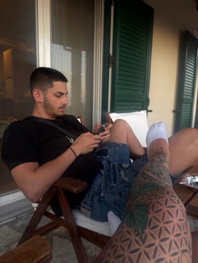 A medium shot depicts two individuals seated on wooden chairs outside a building within the city of Municipal Unit of Lefkada, Greece. One individual is a male with dark hair and a beard, wearing a black t-shirt. He is holding a smartphone. The other person, wearing a pair of distressed denim shorts and socks, is heavily tattooed, and their leg is partially visible in the frame. Shutters are visible in the background.