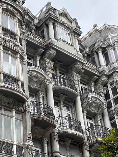The image displays the ornate facade of a historic building in Oviedo, Spain, characterized by its elaborate neoclassical architectural style. The building features multiple stories with curved balconies adorned with intricate wrought-iron railings and detailed stone carvings. These carvings include sculpted figures, floral motifs, and classical architectural elements such as Corinthian columns and pediments. The windows are tall and narrow, with some showcasing sheer white curtains. The stonework appears weathered, suggesting age and exposure to the elements. The sky is overcast, providing a diffused, even light that highlights the textures and details of the facade. A few branches of lush green foliage are visible in the lower right corner, contrasting with the gray stone. The perspective is a low-angle shot, emphasizing the grandeur and verticality of the building.