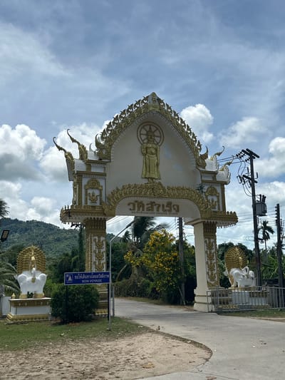 The image shows a large, ornate archway, likely a temple entrance, in Baan Harn, Thailand. The archway is decorated with gold accents and features a statue of Buddha at the apex, along with intricate carvings. Flanking the entrance are two large white statues of mythical creatures, possibly griffins or lions, also adorned with gold. A road leads through the archway, with lush green hills and palm trees in the background. A blue sign with text in Thai and English reads "BON VOYAGE" and is positioned to the left of the entrance. Electrical poles and wires are visible on the right side of the frame, indicating the presence of infrastructure. The sky is partly cloudy with a mix of sun and shade. The overall scene is a snapshot of a cultural and potentially spiritual landmark in a tropical setting.
