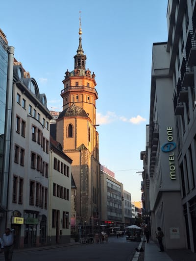 The image captures a street scene in Leipzig, Germany, with a prominent church tower dominating the skyline. The tower, a historical landmark, is bathed in the warm light of the setting sun, casting long shadows on the cobblestone street. Modern buildings with glass facades line the street, creating a contrast between old and new architecture.
Several people are visible in the scene. On the left side of the street, a man in a checkered shirt is walking away from the camera. Further down the street, groups of people are seen dining at outdoor cafes and browsing in shops. On the right side, a lone figure stands on the sidewalk, playing a musical instrument. The street is clean and well-maintained, with parked bicycles and streetlights adding to the urban ambiance. The sky is clear blue, with a few wispy clouds adding texture to the scene. A hotel sign, "MOTEL ONE," is visible on one of the modern buildings. The overall impression is one of a vibrant city that blends its rich history with modern development.