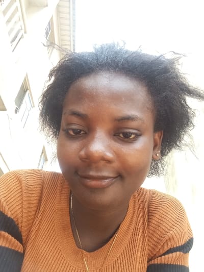 The image displays a low-angle, head-and-shoulders portrait of a young adult female, captured outdoors in what is stated to be Agburu Uke, Nigeria. The subject has dark skin, dark, natural curly hair that appears somewhat disheveled, and is looking slightly downwards with a subtle, closed-mouth smile. She wears small, round stud earrings in both earlobes and a thin, gold-colored chain necklace. Her attire includes a ribbed, rust-orange long-sleeved top with distinct black horizontal stripes on the upper arms. The background is brightly illuminated, showing a light-colored multi-story building featuring visible windows and an upper structural element, possibly a roofline or eave. No other individuals or direct interactions are visible.