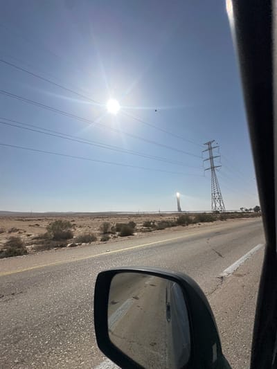 A view from a car window shows a desert landscape with a road, electricity pylons, and a bright, sunlit sky. The car's side mirror reflects the road and landscape. In the distance, there's a concentrated solar power tower reflecting sunlight. The sky is clear blue, with the sun causing a significant glare. The road appears dry and stretches far into the horizon.