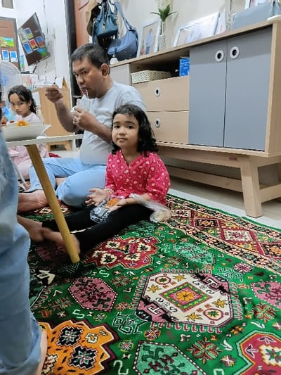 This image depicts an indoor scene in Meunasahbaet, Indonesia, featuring three individuals and part of a fourth, seated on a green geometric patterned rug. A male adult, wearing a white t-shirt and light blue pants, is seated centrally, holding a clear glass and spoon to his mouth, consuming a liquid. To his right and in the foreground, a young female child with dark, curly hair wears a pink patterned long-sleeved top and black pants. She is seated facing the camera, holding a clear plastic bag containing a light-colored item. To the left of the adult male, another young female child, wearing a pink top, is partially visible, seated near a low, light-colored table with a white bowl on it. The lower leg and foot of a fourth individual, wearing light blue pants, are visible in the extreme foreground, suggesting they are also seated on the rug, facing away from the camera. The background reveals a light wood and grey storage cabinet with multiple drawers and doors, adorned with framed pictures, vases, and a white woven basket. Above the cabinet, two blue bags hang from hooks. A wall-mounted television displaying multiple colorful images and a white electric fan are visible on the left side of the frame.