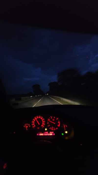 The image depicts a nighttime scene from inside a car, possibly in transit. The car's dashboard, with illuminated red gauges and a digital display showing "Cons: 11.0 L/100", is visible on the right. The view through the windshield shows a dark road with faint markings and distant lights, flanked by trees or foliage silhouetted against a dark blue, cloud-filled sky. The location is not discernable from the image.