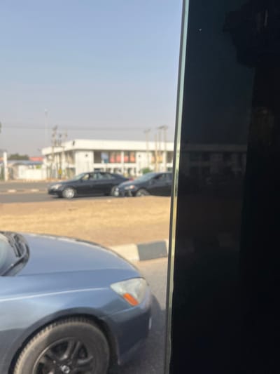 This image captures an urban street scene in the city of Aso, Nigeria, under clear daylight conditions. In the immediate foreground, the front-right quarter of a blue-grey sedan is visible, showing a black-rimmed wheel and its headlight, positioned next to a black-and-white striped curb. Across a median strip of dry, light-brown vegetation, a multi-lane paved road extends into the midground. Two dark-colored sedans are present on this road, one following the other, suggesting active vehicular traffic. In the background, a prominent white two-story building with a flat roof and multiple windows, indicative of a commercial or public structure, stands against a clear, light blue sky. To its left, a smaller white building is partially visible, along with utility poles or power lines. A dark, reflective vertical surface frames the right side of the image, likely an interior panel of the vehicle from which the photograph was taken. No individuals are clearly discernible, nor are any specific actions or interactions taking place beyond vehicular presence.