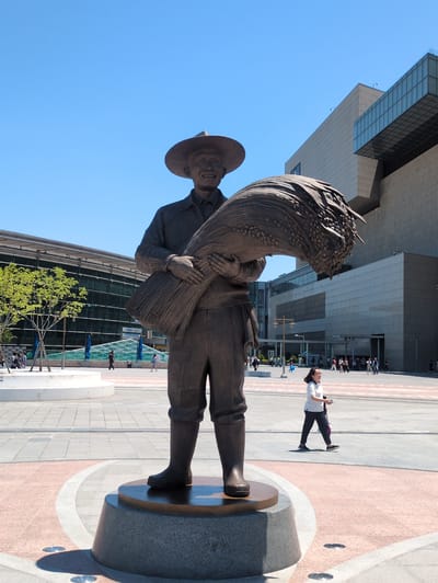 The image shows a bronze statue of a farmer in Daegu, South Korea. The statue is positioned on a round base in a plaza. The farmer is depicted wearing a hat and traditional clothing, carrying a large bundle of grain on his back. A woman is lying on the ground taking a photo of the statue, while a group of people are standing in the background. Tall modern buildings surround the plaza, and the sky is blue.
