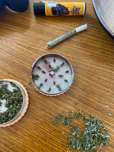 The image presents a top-down view of items laid out on a light-brown wooden surface, likely a table. The scene suggests the preparation of cannabis for consumption.
In the center-left of the frame, there is a pre-rolled item, consistent in appearance with a cannabis joint. It has a white, cylindrical filter tip on one end and is wrapped in translucent rolling paper, allowing the light-green, finely ground plant material inside to be visible. The joint is positioned diagonally, pointing towards the upper right.
Immediately to the left of the joint is a yellow Clipper brand lighter. The lighter has a black cap and base. Text on the lighter, oriented upside down, reads "LION ROLLING CIRCUS" above a graphic of what appears to be a lion tamer or performer. Below the graphic, "CLIPPER" is clearly visible, and underneath that, the word "reusable" can be seen.
Below the joint, in the central part of the image, lies the top section of a circular cannabis grinder. This grinder features a silver-colored metal interior with numerous small, diamond-shaped grinding teeth arranged in a circular pattern. Bits of green, ground plant material are scattered among these teeth and on the metal surface. The grinder's outer rim is a light, possibly beige or off-white, material with a decorative scalloped edge.
Further down and slightly to the right, the bottom section of a similar circular grinder is visible. This section is filled with a dense layer of finely ground green plant material, covering the internal surface completely. It also has the same light-colored, scalloped outer rim as the top section of the other grinder.
In the upper right portion of the image, a loose pile of finely ground green plant material, consistent with cannabis, is scattered directly on the wooden surface. This material appears similar in texture and color to what is seen inside the pre-rolled joint and in the bottom grinder section.
The overall impression is one of an ongoing process, with plant material having been ground and some already rolled into a joint, suggesting preparation for smoking. The wooden surface shows a distinct grain pattern, running mostly horizontally. The lighting is bright and even, indicating an indoor setting, possibly during the day.
