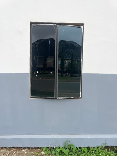This media file displays a close-up view of a wall painted in two distinct colors: white on the left and a lighter shade of gray on the right. Embedded within the wall is a rectangular window with dark, reflective glass panes. The window is divided horizontally, with two panes stacked vertically, creating a grid-like appearance. The reflections in the glass reveal a modern building with multiple floors and a facade that appears to be composed of glass panels and possibly solar panels. Green foliage, likely trees or bushes, is also visible in the reflection, suggesting an outdoor environment.
To the right of the gray section of the wall, a small patch of green vegetation grows alongside the wall, with small white flowers interspersed among the leaves. The ground beneath the vegetation is composed of dirt and small stones. The overall scene suggests an exterior wall of a building, possibly in an urban or suburban setting, given the reflection of the multi-story building. The lighting in the image indicates daylight, likely midday, with the sun casting a bright illumination on the wall. There is no discernible text in the image. The location context is identified as Karum, Nigeria.