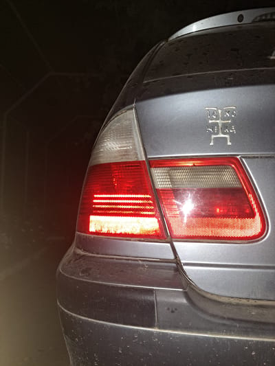 The image shows the rear left side of a silver-colored car at night, parked in what appears to be an alleyway or passage in the city of Vypasne, Ukraine. The car's left taillight is illuminated, casting a red glow. The taillight assembly is partially visible, showing red and clear sections. The car's body appears to be dusty and has some minor scratches. Above the taillight, on the trunk lid, there is a silver emblem with Cyrillic letters that read "IXC NIKA". The background is dark and out of focus, suggesting a dimly lit environment. The ground surface appears to be asphalt or concrete. There are no people visible in the image.