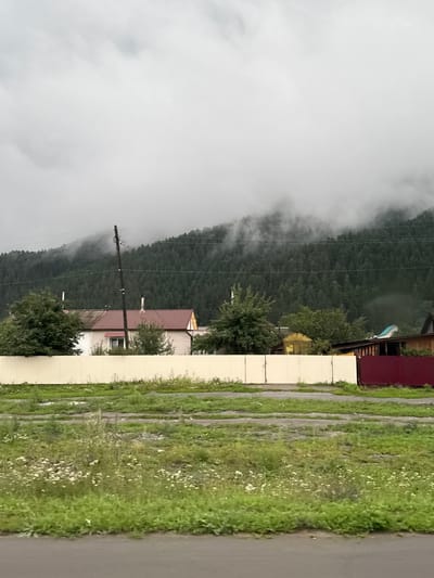 The image depicts a rural scene in Elekmonar, Russia, characterized by a overcast sky and a forested hillside partially shrouded in mist. In the foreground, a beige fence runs horizontally across the frame, separating a grassy area with wildflowers from what appears to be a dirt or gravel road. Beyond the fence, several buildings are visible. One structure has a light-colored exterior with a prominent red roof and a single visible window. A utility pole stands near this building. Further to the right, a wooden structure with a dark red fence is partially visible, along with a smaller yellow greenhouse-like structure. The dense green forest dominates the background, climbing the hillside and disappearing into the clouds and fog. The overall atmosphere is subdued and tranquil, suggesting a cool or damp day.