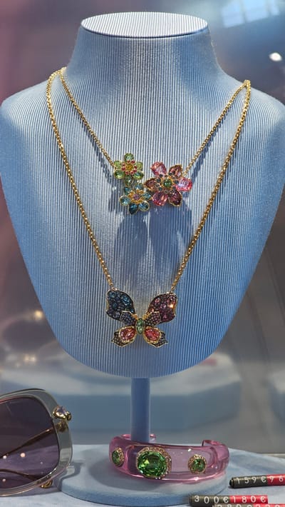 A close-up, horizontal shot showcases a display of jewelry on a light blue mannequin bust. The primary subject is a delicate gold chain necklace adorned with two charms: a cluster of three colorful gemstone flowers and a sparkling butterfly. The flowers are rendered in shades of pink, green, and blue, with small clear stones accenting their petals. The butterfly charm is equally vibrant, featuring wings in purple, pink, and green, also embellished with small, clear gemstones.

To the right of the necklace, a pink, translucent bangle bracelet is displayed. This bracelet is encrusted with large green gemstones and smaller clear stones. Below the bracelet and slightly to the right, a pair of sunglasses with a silver frame and purple lenses are partially visible, featuring a small heart-shaped charm on the right temple.

The background is softly blurred, suggesting a display window or a retail environment. The lighting appears to be bright, possibly from an indoor source or sunlight, casting gentle shadows. The scene is likely a jewelry store in Zaragoza, Spain, given the context.

Visible text includes price tags to the right of the pink bangle, partially obscured but showing numbers like "159 €99" and "3000 €180 €". There is also faint text on the sunglasses, possibly a brand name. The overall impression is one of elegance and luxury, highlighting the intricate craftsmanship of the displayed jewelry. The focus is entirely on the merchandise, with no people present in the frame.