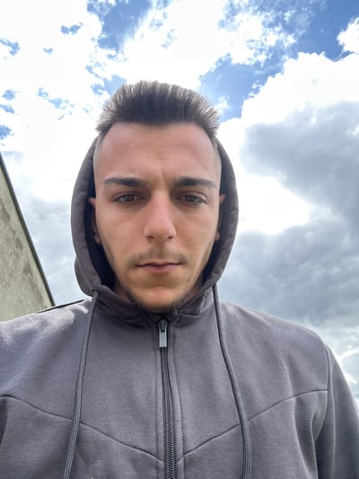 A low-angle, close-up selfie features a male individual wearing a gray hooded sweatshirt with the hood up. The zipper of the sweatshirt is partially visible. The man has short, dark hair, brown eyes, and light facial hair. The background consists of a cloudy sky with patches of blue. The location is Srednia, Bulgaria. There is a light-colored building in the very lower portion of the picture.