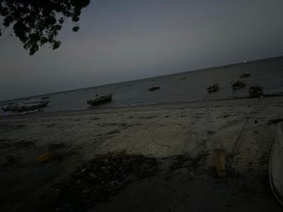 The image presents a dark, coastal scene at late dusk or early night, likely in Bagamoyo, Tanzania. The sky is uniformly dark gray and overcast, casting a somber light over the landscape.
In the foreground, a sandy and muddy beach occupies the lower half of the frame. Towards the bottom center, there is an accumulation of dark debris or trash, and to its left, a bright yellow bucket or container is visible on the sand. A thick wooden post stands upright in the sand to the right of the debris, with a rope extending towards the right edge of the frame. Part of a white boat hull is cropped at the far right.
Numerous traditional wooden boats are present across the scene. On the far left, one boat, partially beached, shows a faint blue light or reflection. Another boat rests closer to the water's edge towards the mid-left. Several more boats are afloat in the calm, dark water, particularly on the right side of the image where they appear clustered. The water itself is dark and still, reflecting the dim ambient light.
Dark, silhouetted tree branches with sparse leaves overhang the scene from the top left corner. The overall atmosphere is quiet and still, suggesting a peaceful end to the day in a working coastal community. No people are visible, and no discernible text appears anywhere in the image.