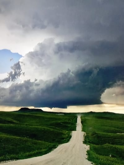 The image depicts a rural landscape dominated by a dramatic sky filled with dark, ominous storm clouds. A winding dirt road cuts through rolling green hills, leading the viewer towards a distant, flat-topped hill. The overall atmosphere is one of impending weather, with the light filtering through the clouds suggesting either dawn or dusk. The scene is entirely devoid of human presence or any signs of urban development. The provided crops only show close-ups of the sky and the surrounding terrain, reinforcing the focus on the natural elements. There is no information that allows for the identification of a specific location, let alone a city like Chachersk, Belarus.