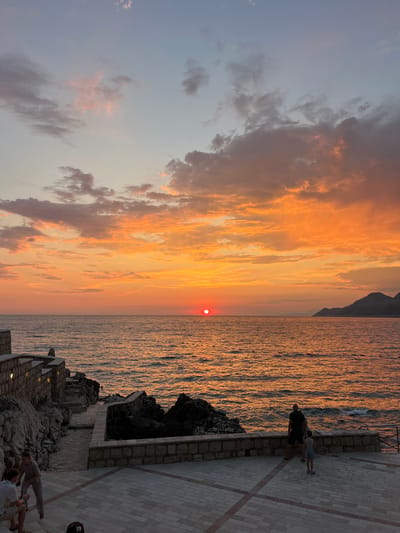 A vertical, full shot captures a sunset over the Adriatic Sea near Pečurice, Montenegro. The sky is ablaze with fiery orange and pink clouds, streaked with muted grays and blues. The sun, a vibrant red disc, hovers just above the horizon. The sea reflects the sky's warm hues, its surface rippling.
In the foreground, a paved walkway runs along a rocky coastline. Two people, silhouetted against the light, stand near the edge, gazing out at the sea. Further along the walkway, a low stone wall and building structures are visible, integrated into the rocky terrain. The scene suggests a peaceful evening, with the warm colors of the sunset evoking a sense of calm and beauty. The lighting indicates it is late afternoon or early evening.