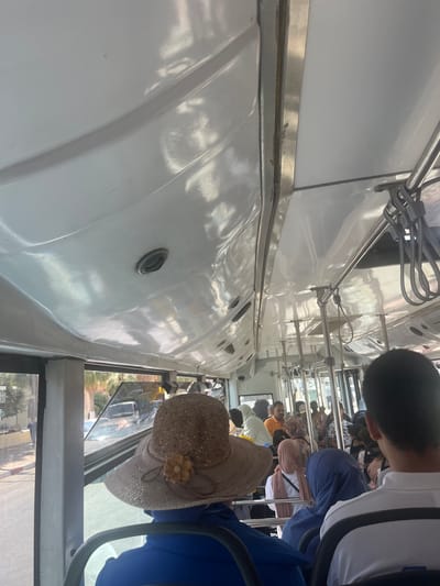 The image shows the interior of a crowded bus in Boufakrane, Morocco. The ceiling of the bus is white and slightly curved, with visible air vents and metal handrails. Several passengers are seated, and some are standing, indicating a full vehicle. One passenger in the foreground wears a wide-brimmed straw hat adorned with a decorative flower. Another passenger in the foreground is wearing a blue headscarf. Outside the windows, glimpses of a street scene can be seen, including buildings, trees, and other vehicles, consistent with an urban or semi-urban environment in Morocco. The overall impression is that of a typical public transportation scene during the day.