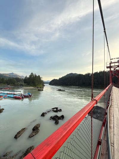 The image shows a view from a suspension bridge in the city of Barangol, Russia. The bridge has a red railing and a wooden walkway. A river flows below the bridge, with boats docked on the left side. Trees line the banks of the river, and mountains are visible in the distance. The sky is partly cloudy, with the sun setting in the background, casting a warm glow on the scene.
