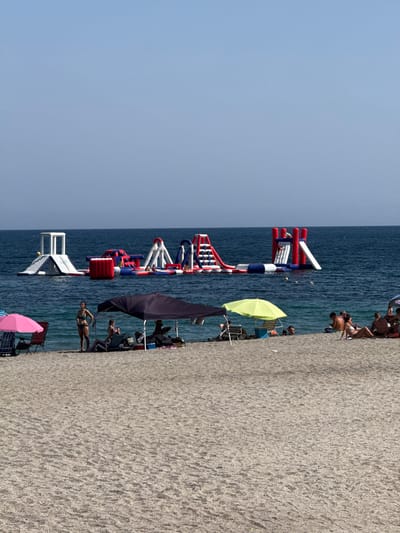 A wide shot captures the sandy beach of Campillo del Moro, Spain, under a clear blue sky. In the foreground, beachgoers relax under umbrellas on the sand. A pink umbrella is visible on the left, and a dark canopy and a bright yellow umbrella offer shade further back. Several people are seated on beach chairs or lounging on towels. In the middle ground, the Mediterranean Sea is calm and a vibrant inflatable water park with slides and climbing structures is set up. Several people are visible on and around the inflatable structures. In the background, the deep blue water stretches to the horizon.