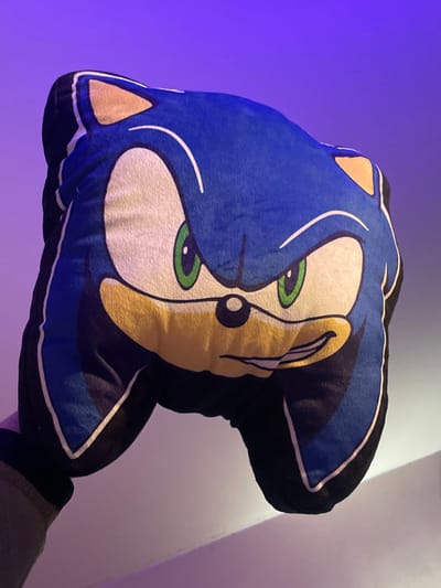 The scene depicts a plush cushion, designed in the likeness of Sonic the Hedgehog's head, held by a human hand. The cushion features Sonic's signature blue fur, a peach-colored muzzle, green eyes with black pupils, and a determined expression with a slight smirk. The hand, visible in the lower left quadrant, is clad in a garment sleeve, appearing to be olive green or beige. The background consists of a plain wall, illuminated by artificial lighting that casts a gradient of purple and magenta hues, transitioning from a deeper purple at the top to a lighter, pinkish-purple towards the bottom. The location of this scene is in the city of Plaisir, France.